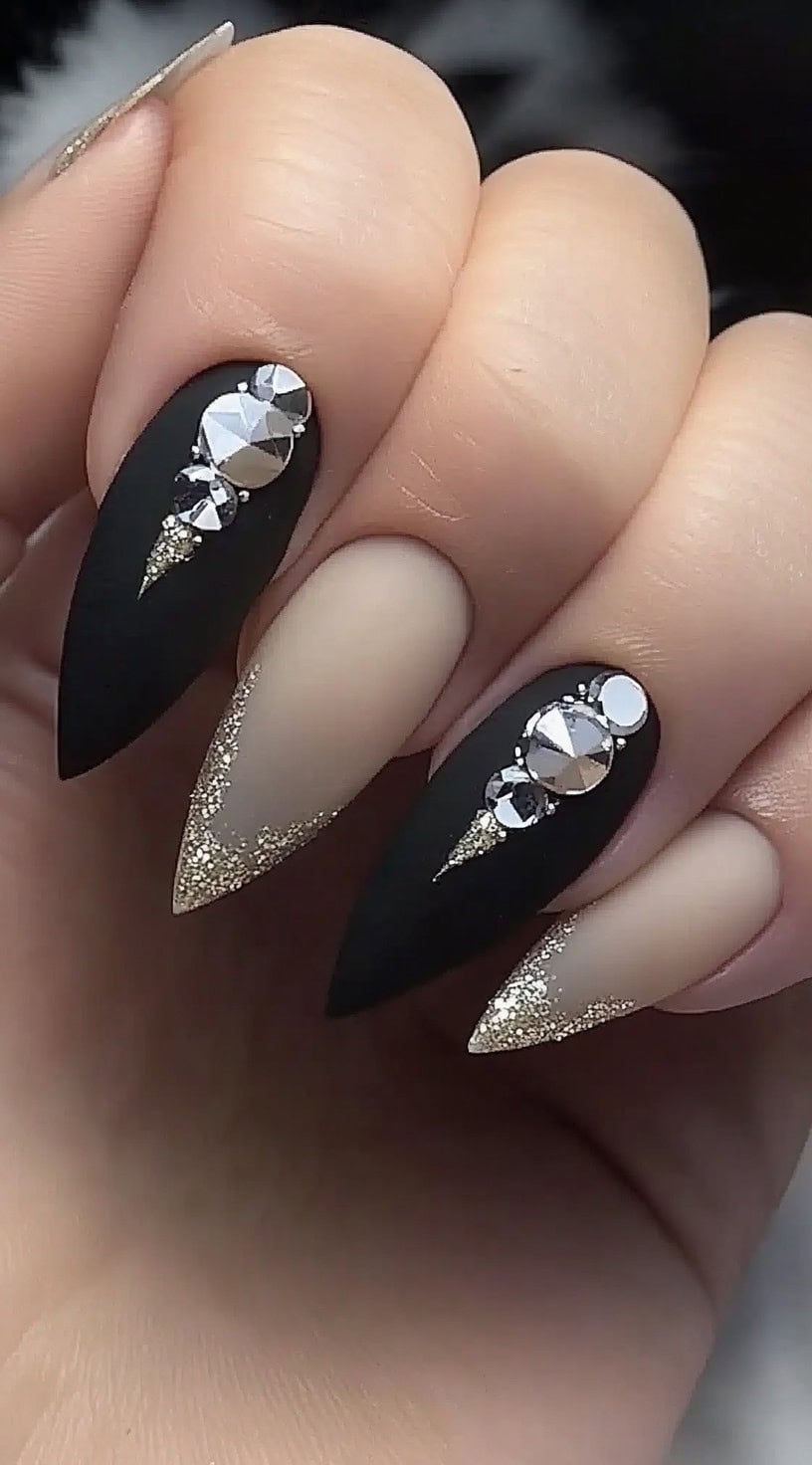 A hand with stiletto-shaped nails featuring a mix of matte black and sheer nude designs, adorned with rhinestones and gold glitter tips.