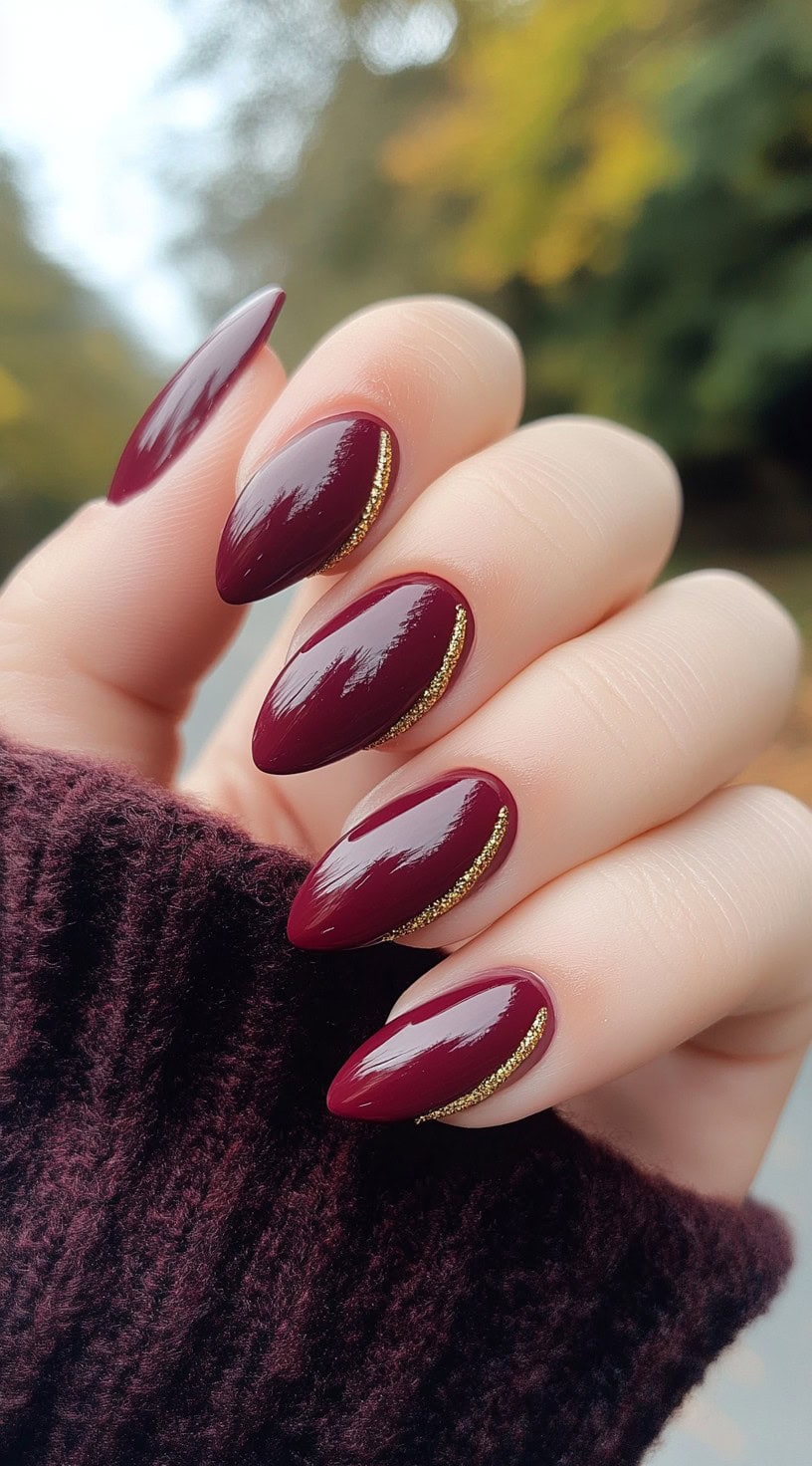 Almond-shaped nails painted in deep burgundy with delicate gold accents lining the cuticles.