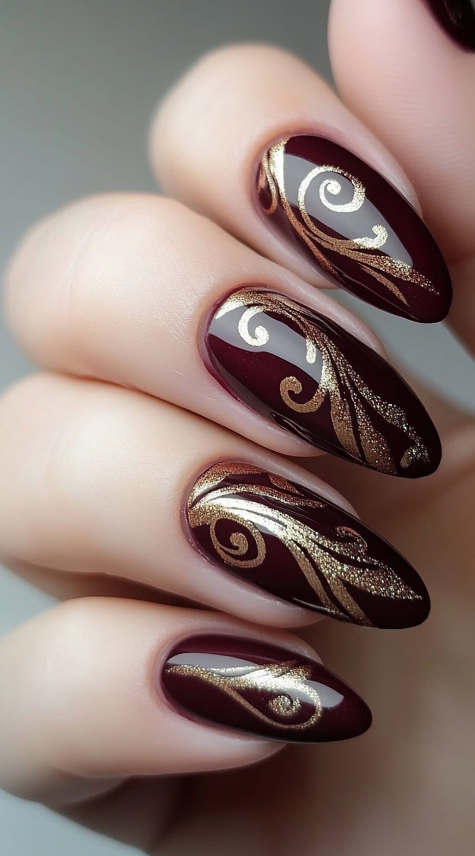 Almond-shaped burgundy nails featuring intricate gold swirl designs for a luxurious effect.
