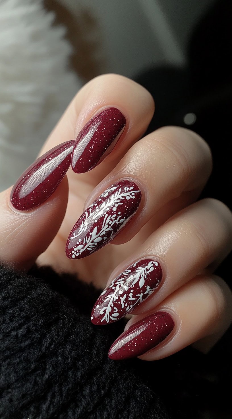 Almond-shaped nails in deep burgundy with delicate white floral detailing and fine glitter for a romantic effect.