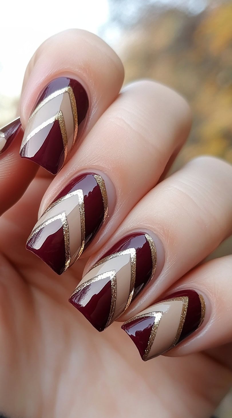 A hand with square-shaped nails featuring a geometric chevron pattern in burgundy, nude, and gold accents.