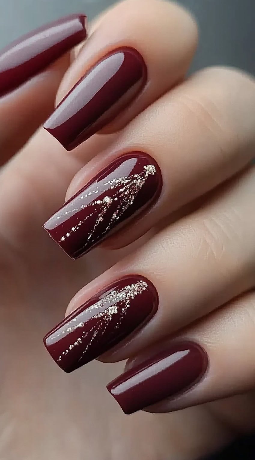 A hand with glossy burgundy nails featuring subtle gold glitter streaks on two accent nails.