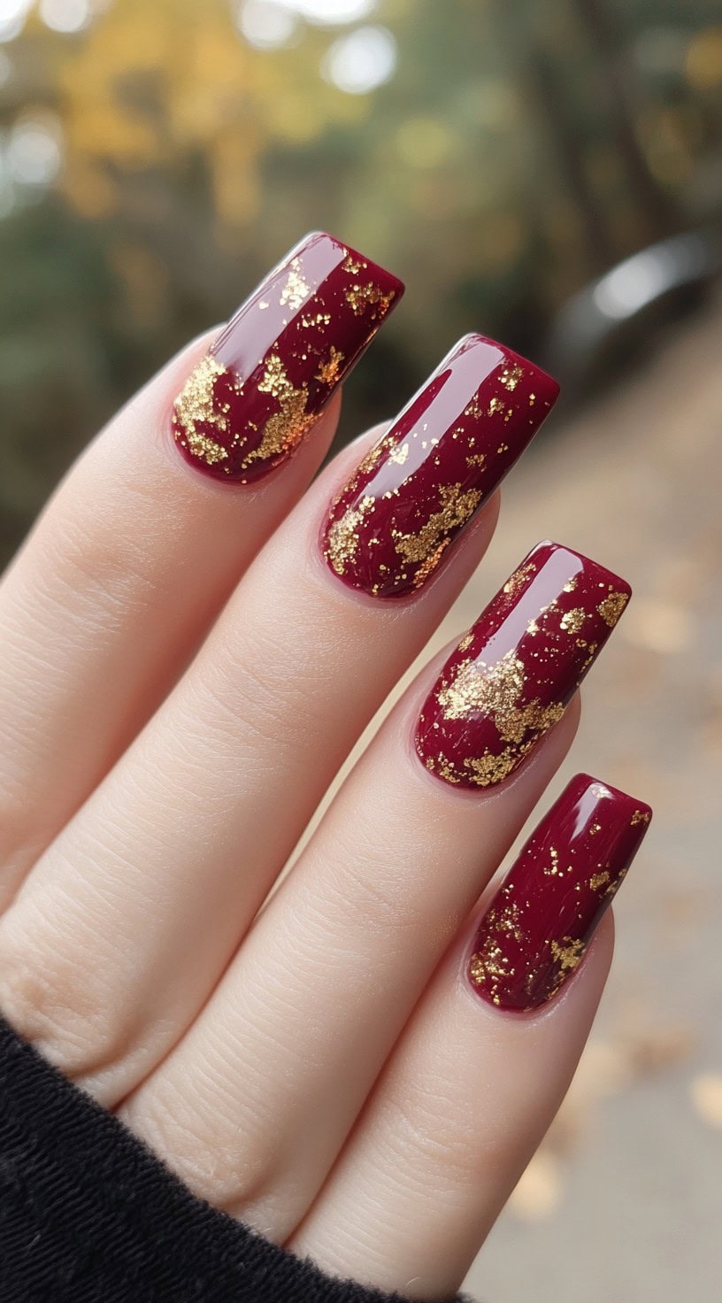 Long square-shaped nails coated in deep burgundy with gold leaf foil embellishments for a luxurious finish.