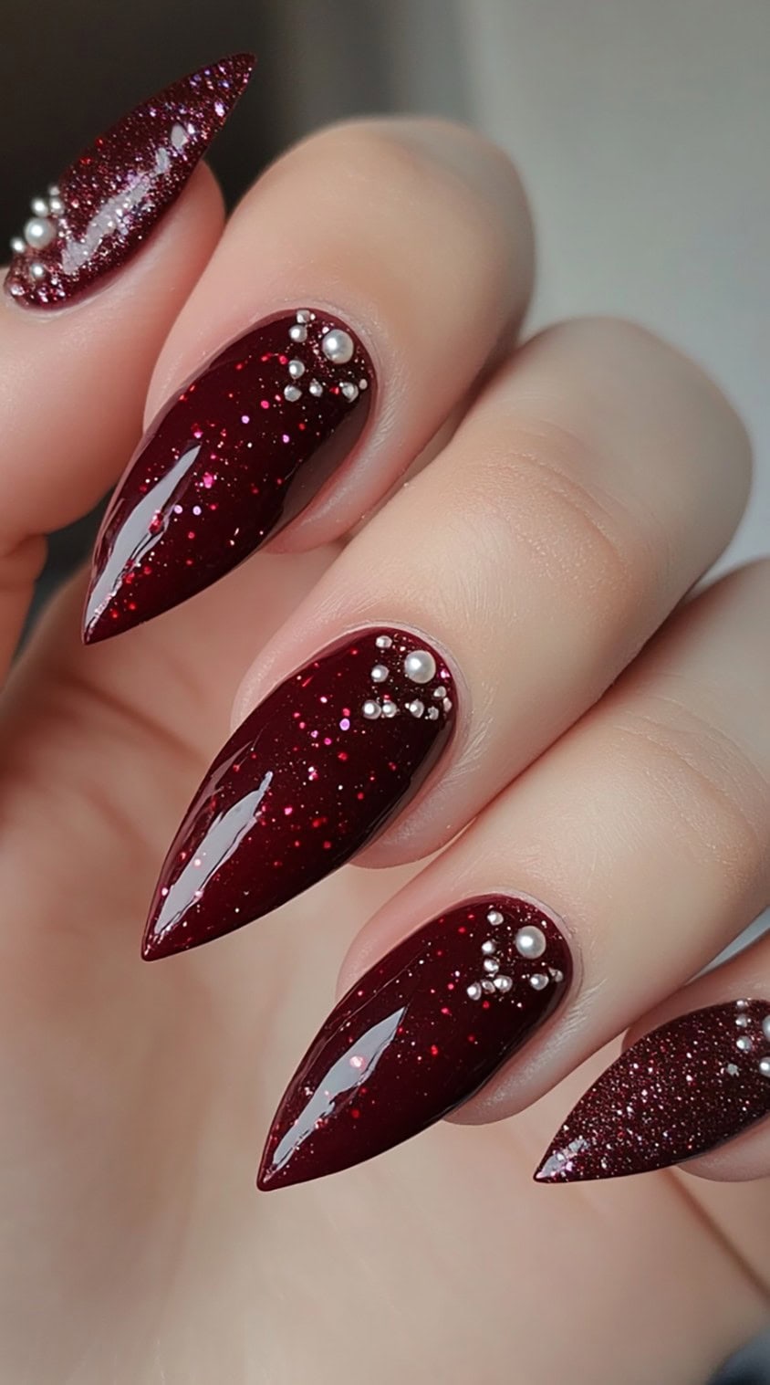 Stiletto-shaped burgundy nails with fine glitter and pearl embellishments on a high-gloss finish.