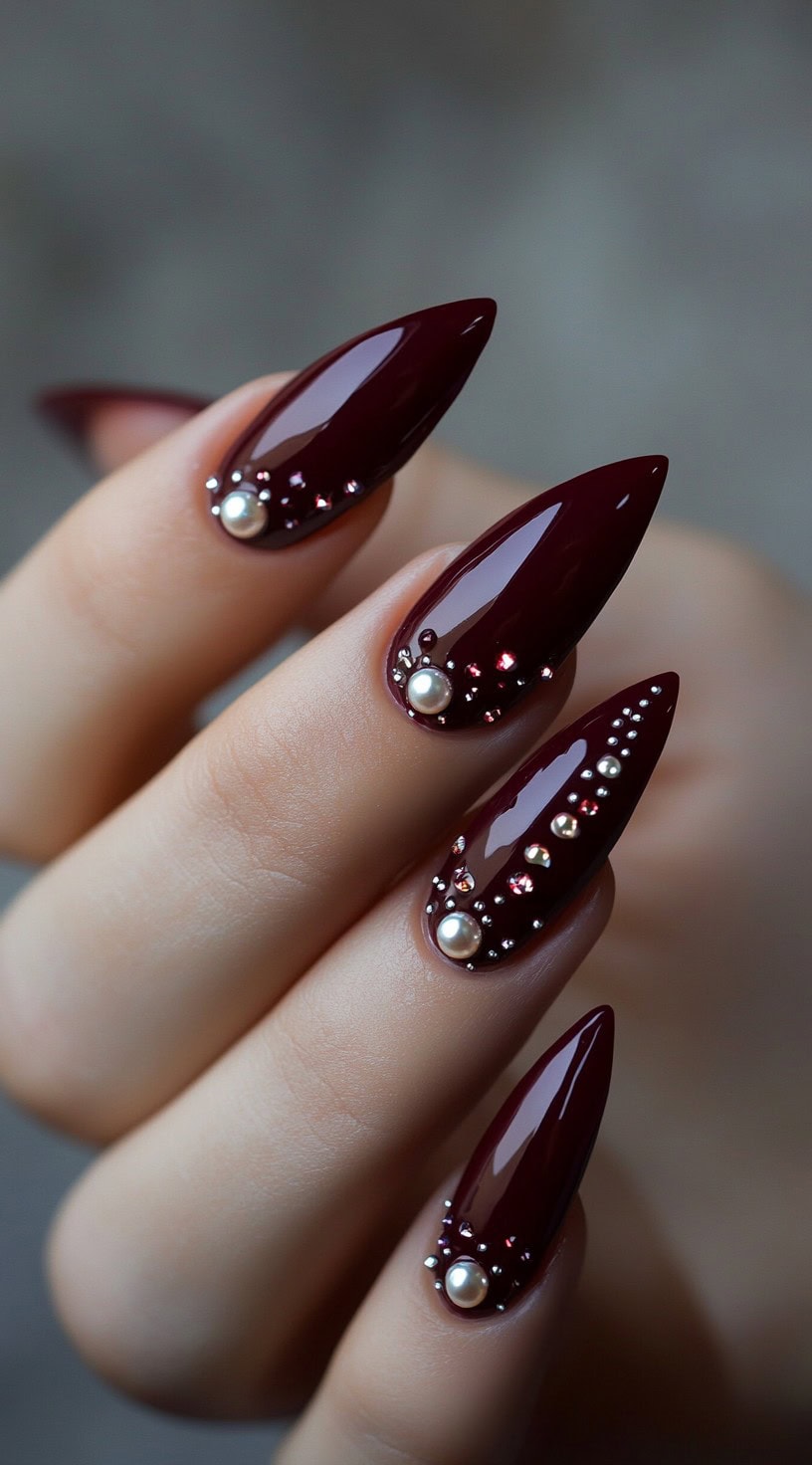 Stiletto-shaped nails with a deep burgundy base, decorated with pearls and tiny crystal embellishments.