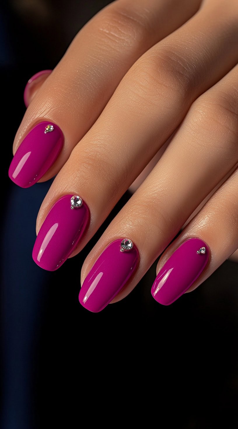 A hand with glossy magenta nails, each featuring a small rhinestone accent near the cuticle, against a dark background.