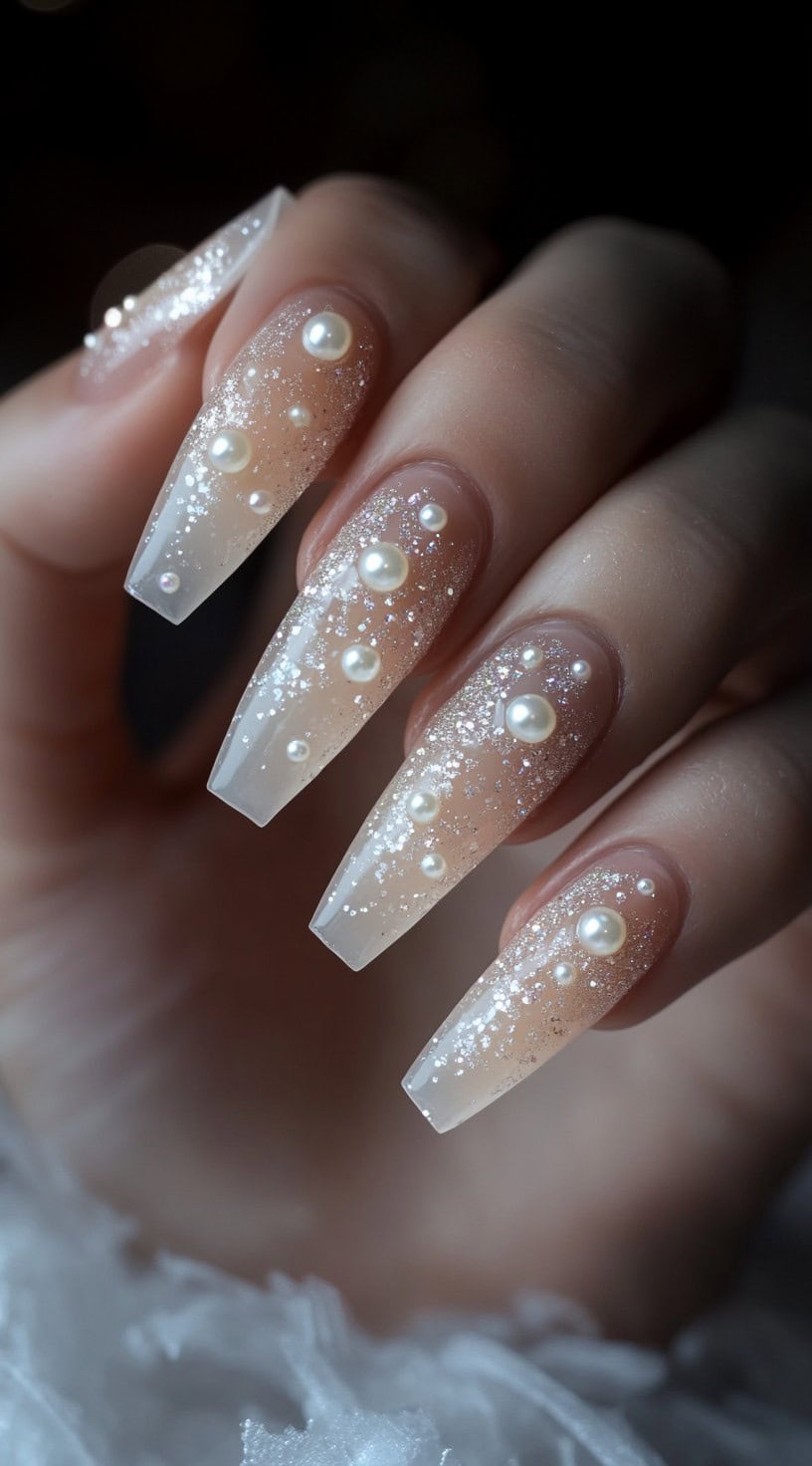 Long coffin-shaped nails with a nude-to-clear gradient, dusted with fine glitter and adorned with scattered pearls.