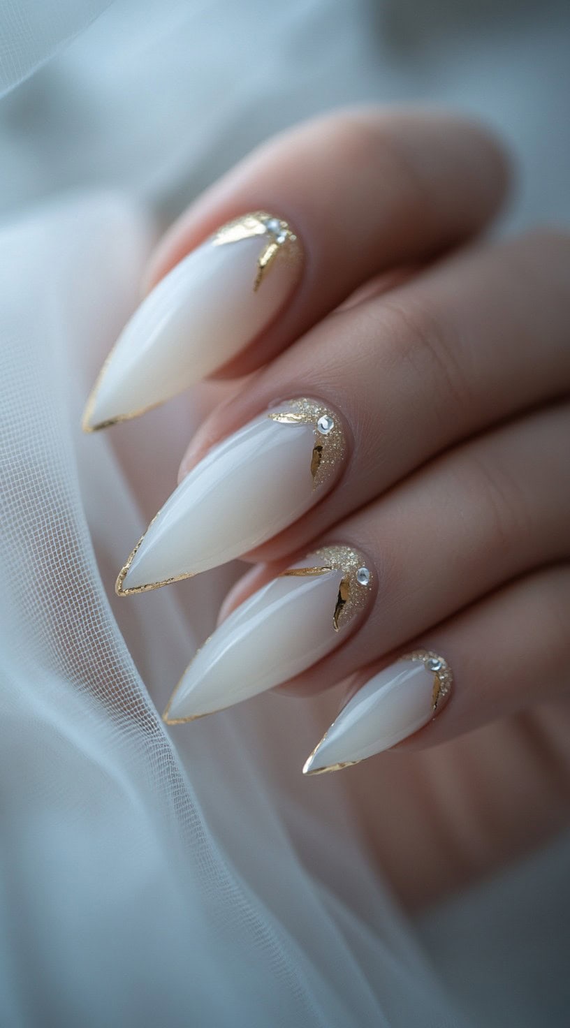 Sharp stiletto-shaped nails with a soft white base, gold foil edges, and crystal accents for an opulent bridal look.
