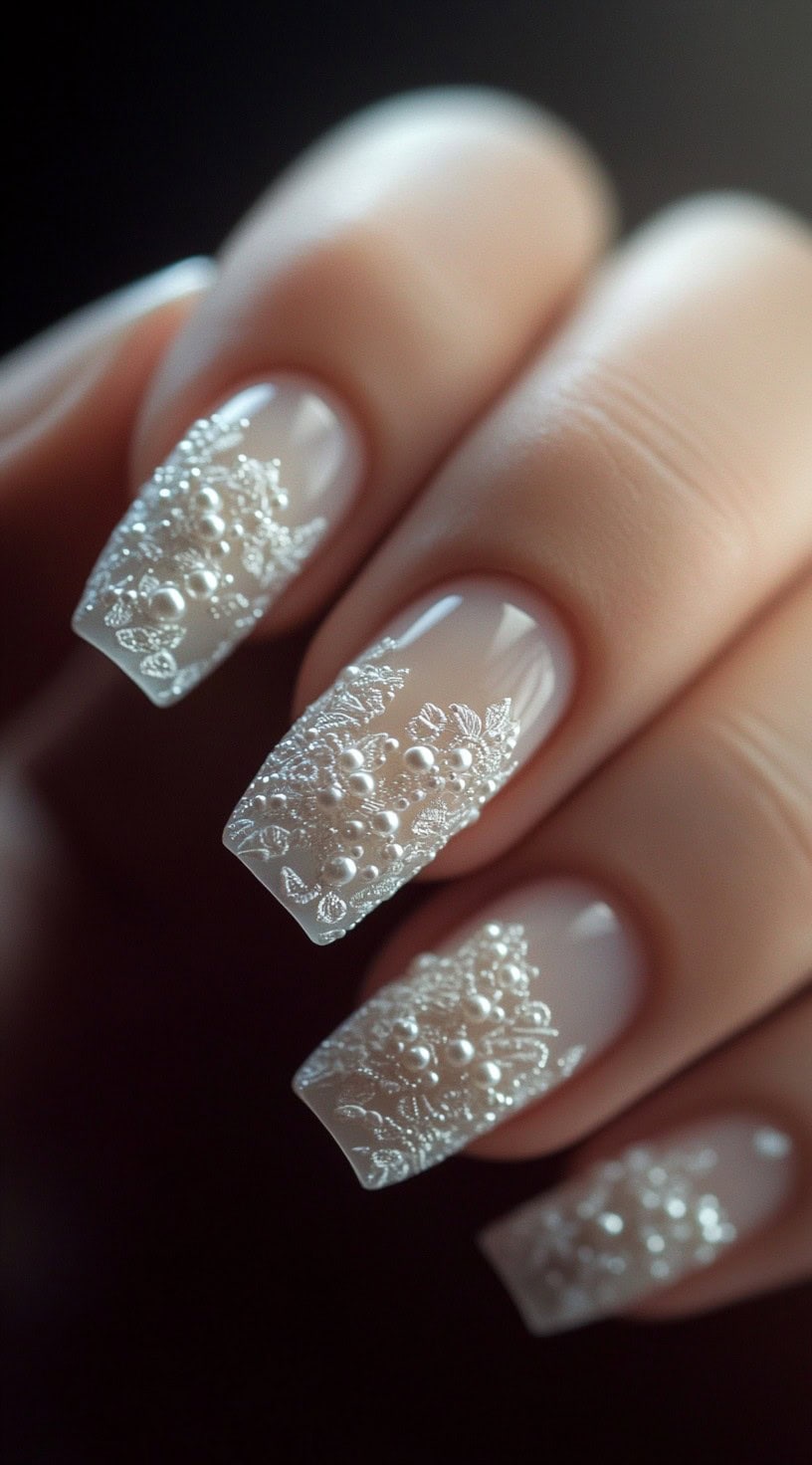 A close-up of square-shaped nude nails adorned with white lace-like 3D embroidery and pearl embellishments.