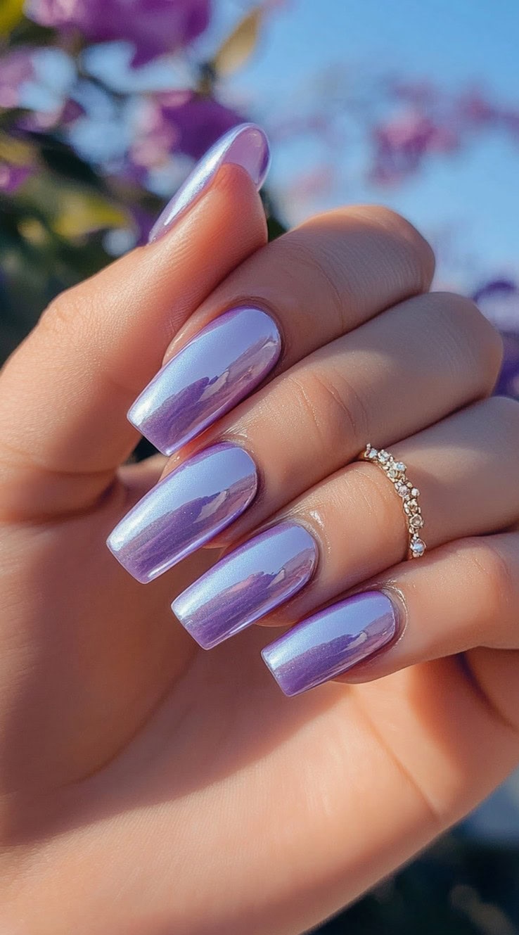 A hand with long coffin-shaped nails featuring a reflective, metallic lavender finish, catching the sunlight against a floral backdrop.