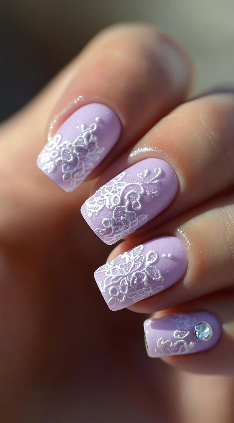 Square-shaped nails painted in soft lilac with embossed white lace details and a rhinestone accent on one nail.