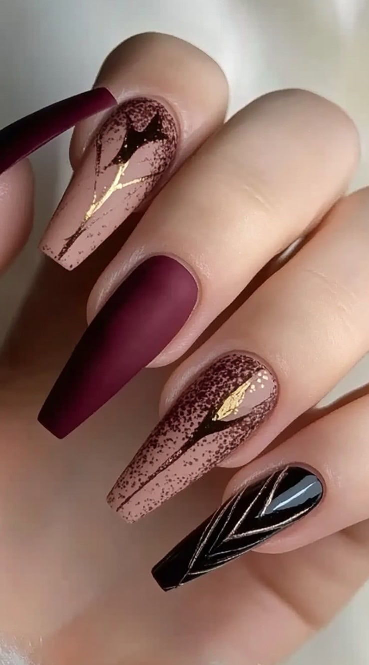 A hand with long coffin-shaped nails in matte burgundy, nude ombré, and gold leaf accents.