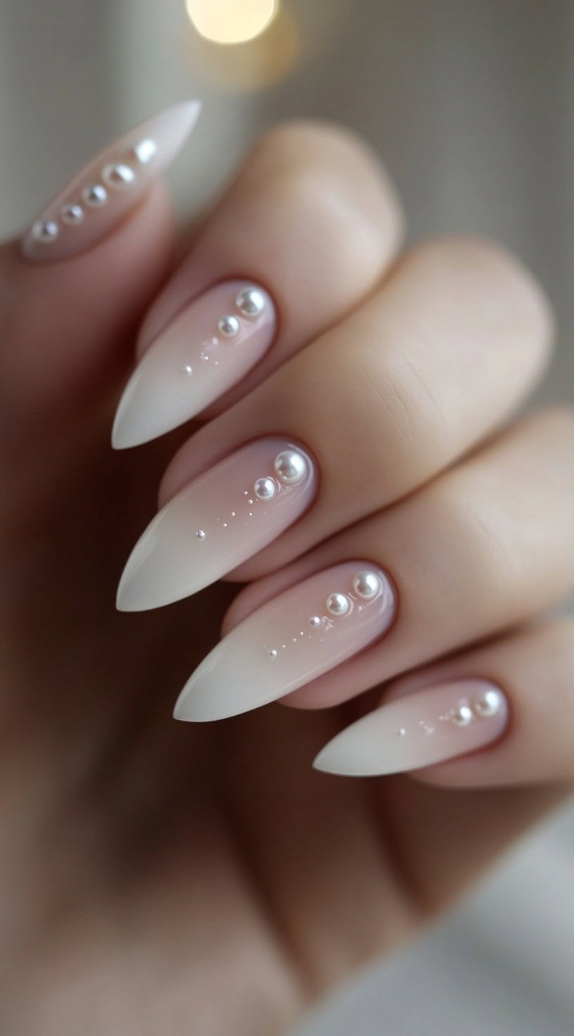 A set of almond-shaped nails with soft white ombre and pearl embellishments placed in a cascading pattern.