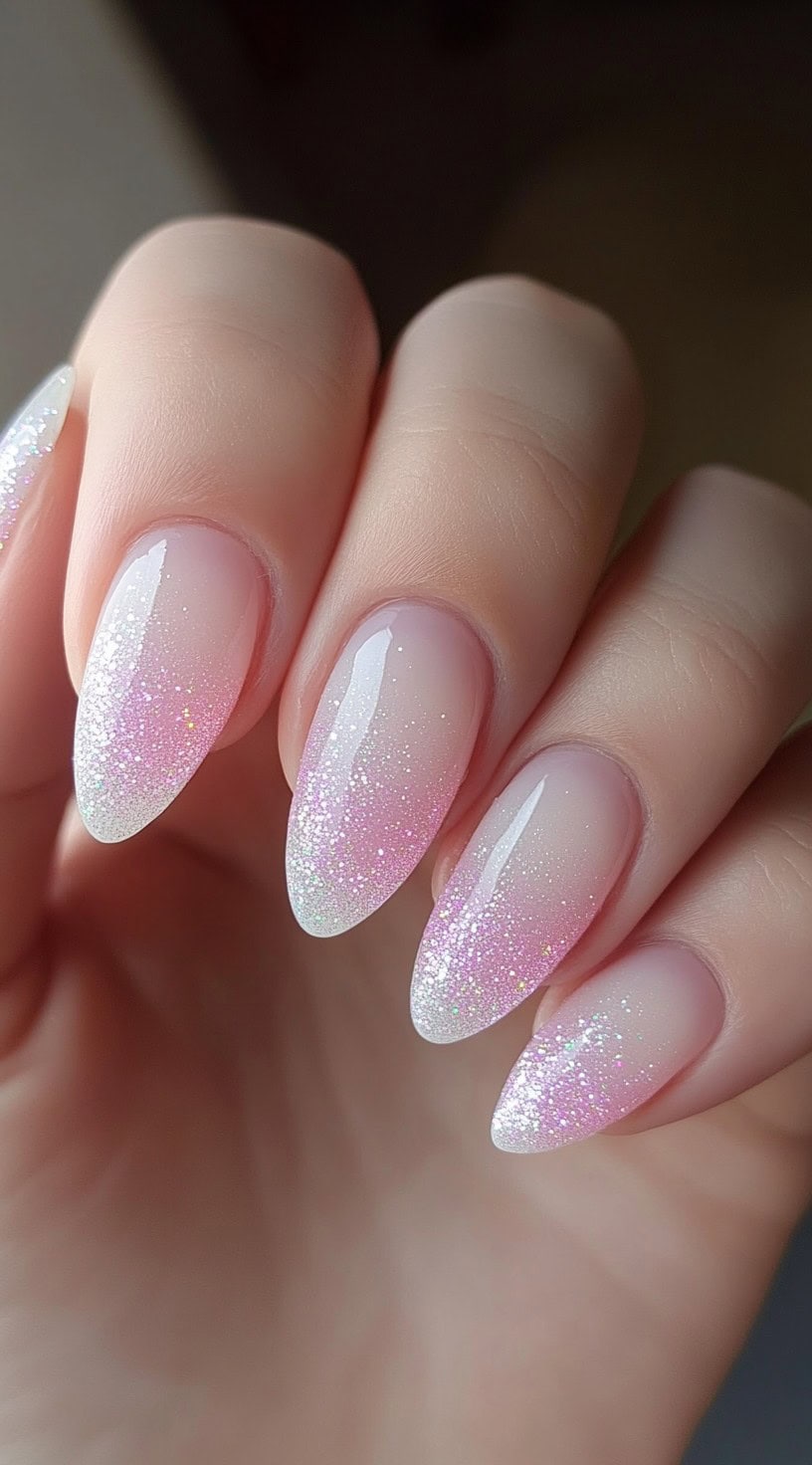 A hand with almond-shaped nails featuring a soft pink and white gradient with ultra-fine iridescent glitter creating a frosty, shimmering effect.
