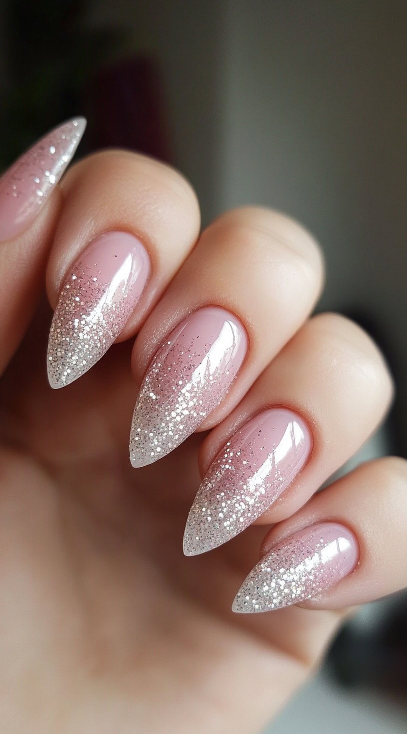 A hand with almond-shaped nails featuring a sheer pink base with a sparkling silver glitter gradient starting from the tips, creating a delicate and ethereal effect.