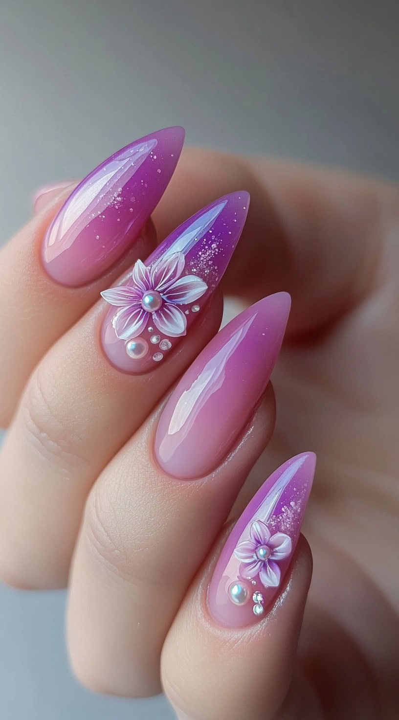 Almond nails featuring a pink-to-purple ombre design with 3D flower accents, pearls, and soft glitter details.