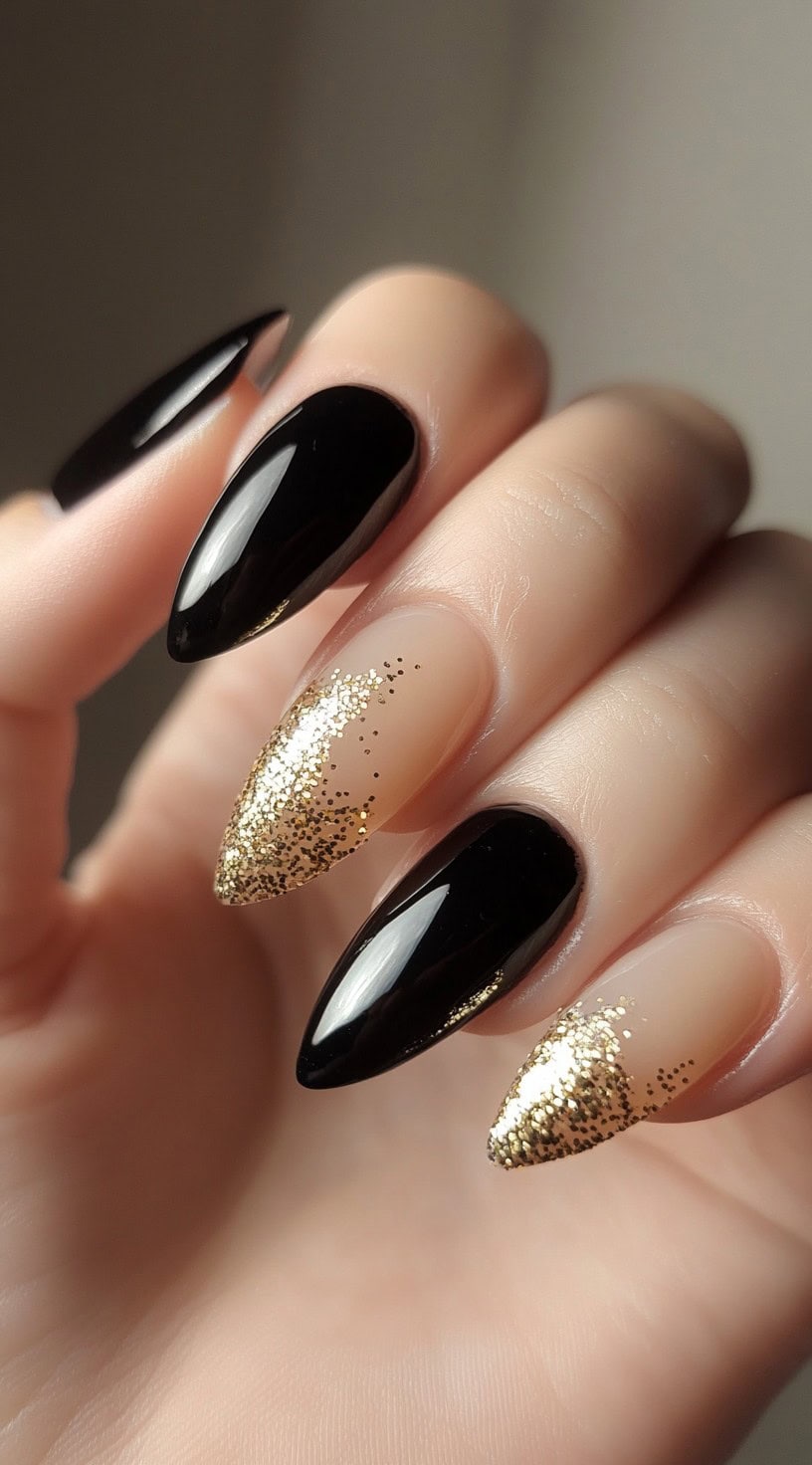 A hand with almond-shaped black nails, featuring a gradient of gold glitter cascading from the tips.