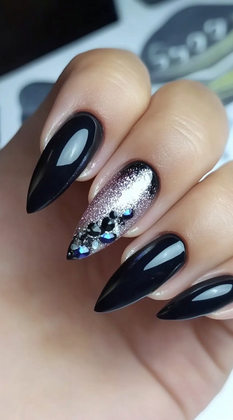 A hand with almond-shaped nails painted glossy black, with one accent nail featuring silver glitter and rhinestone embellishments.