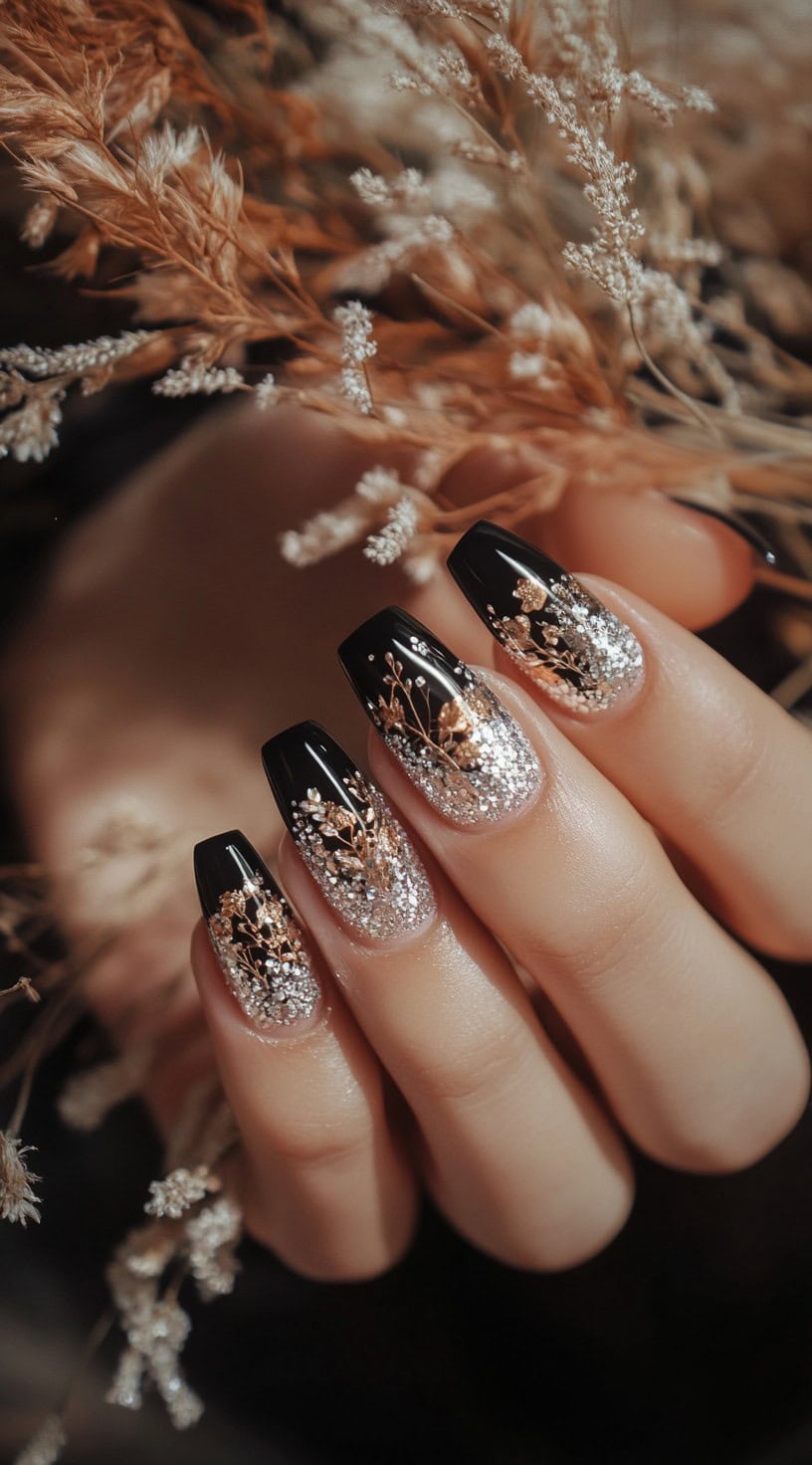 A set of square-shaped nails transitioning from a black base into a silver glitter ombre effect, adorned with delicate metallic floral details.