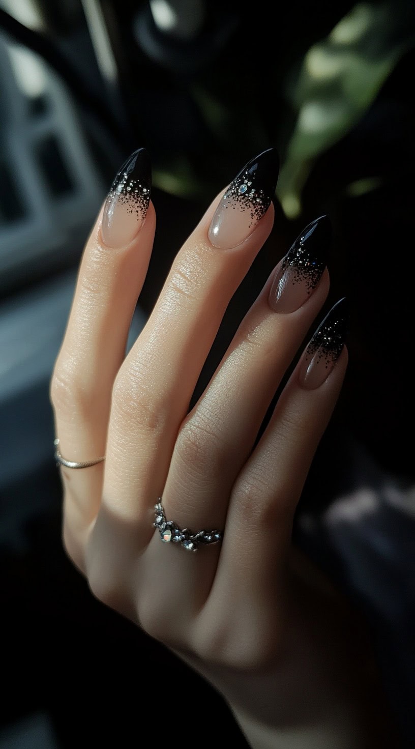 A hand with almond-shaped nails featuring a sheer nude base that transitions into black tips with scattered glitter.