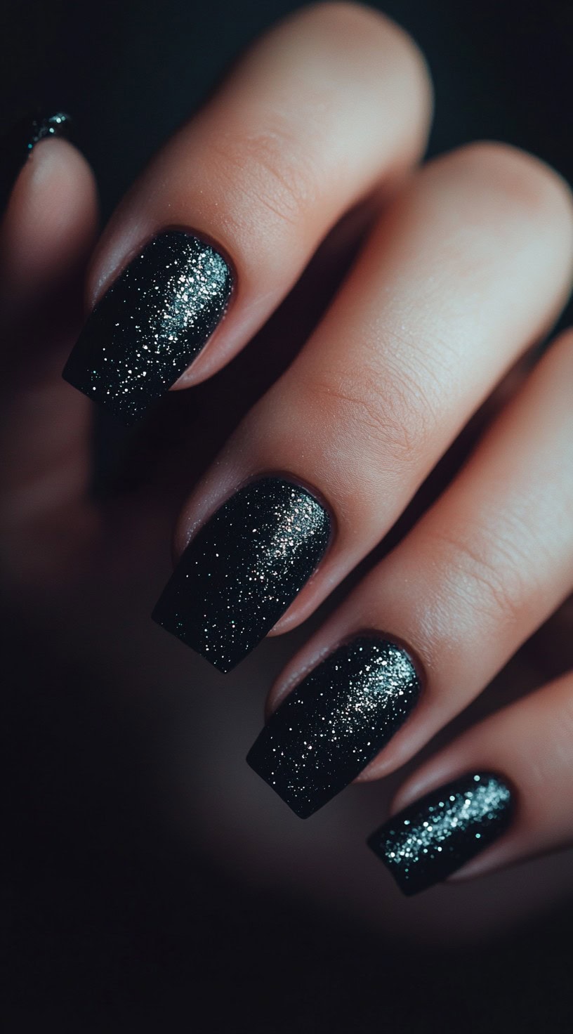A hand with square-shaped nails painted in deep black with an intense glittery finish that sparkles under the light.
