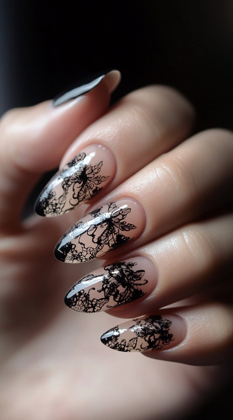 A set of almond-shaped nails with sheer nude polish and intricate black lace patterns extending from the tips.