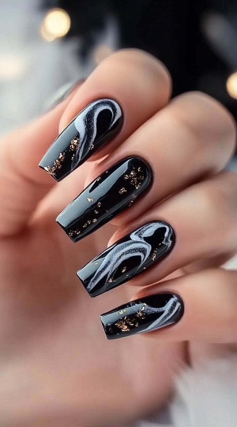 A hand with square-shaped nails painted in glossy black, featuring delicate white marble swirls and gold foil accents.