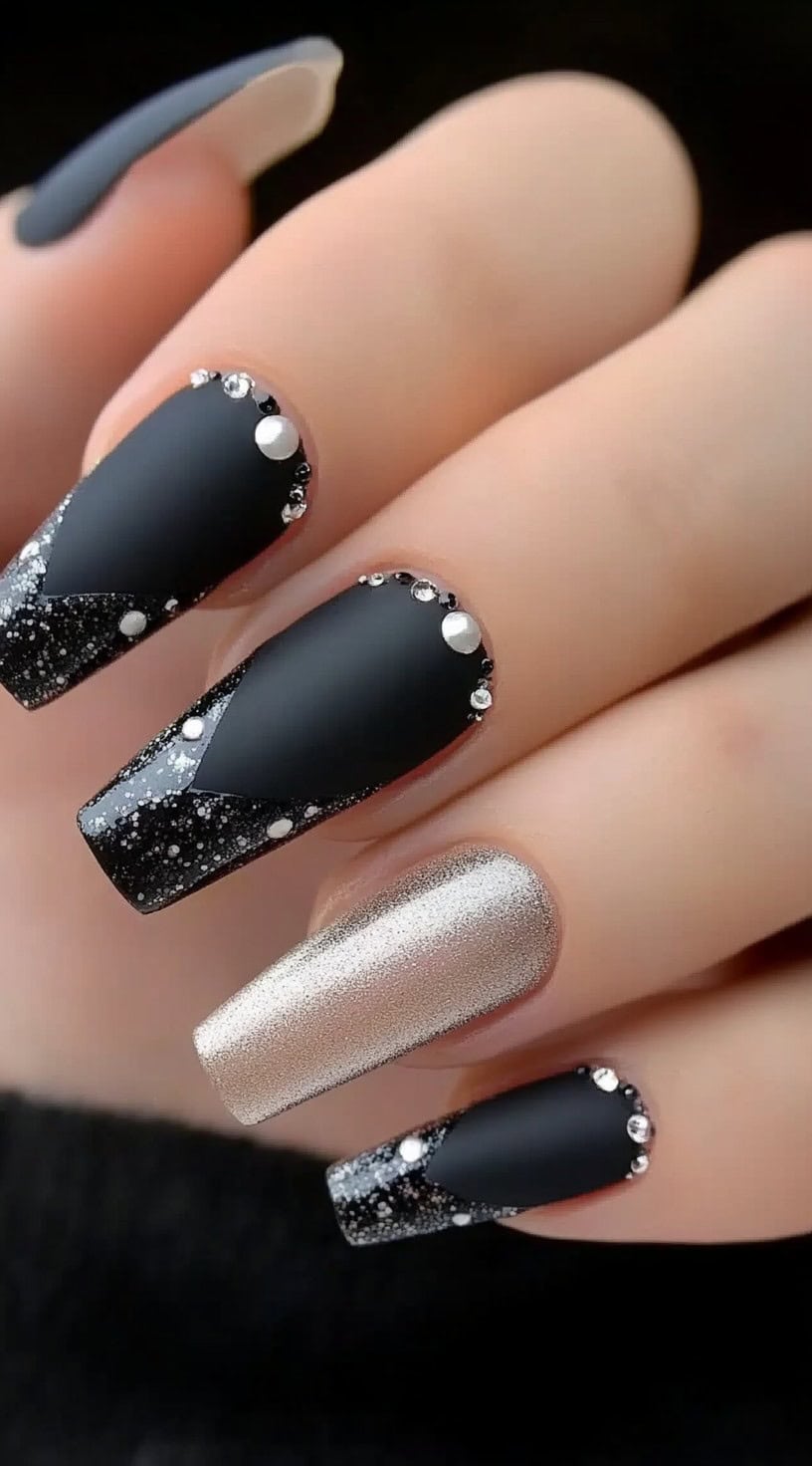 A hand with square-shaped nails featuring a mix of matte black polish, glitter accents, and rhinestones along the nail edges.