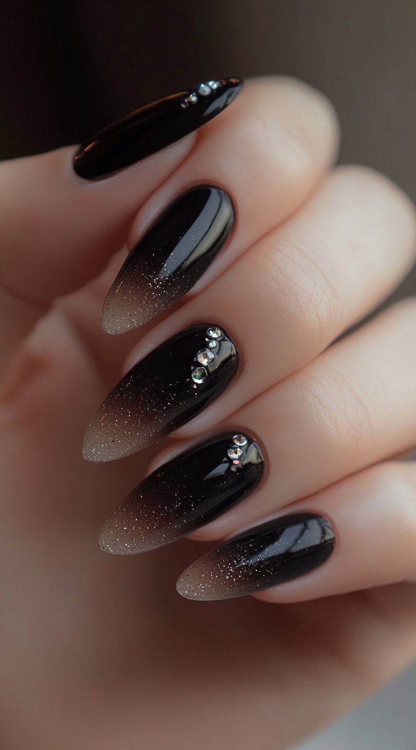 A hand with almond-shaped nails featuring a black-to-nude ombre design with subtle glitter and rhinestone accents on select nails.