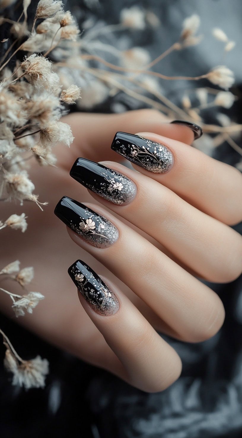 A hand with square-shaped nails featuring a black-to-sheer ombré gradient with delicate floral silver glitter accents.