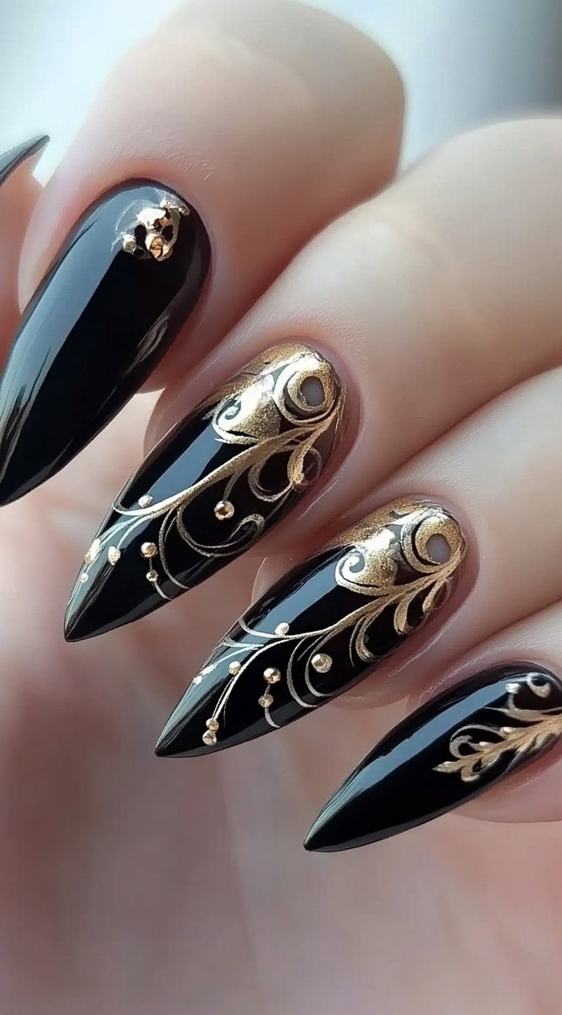 A hand with stiletto-shaped nails painted glossy black, adorned with intricate gold baroque-style patterns and tiny metallic beads.