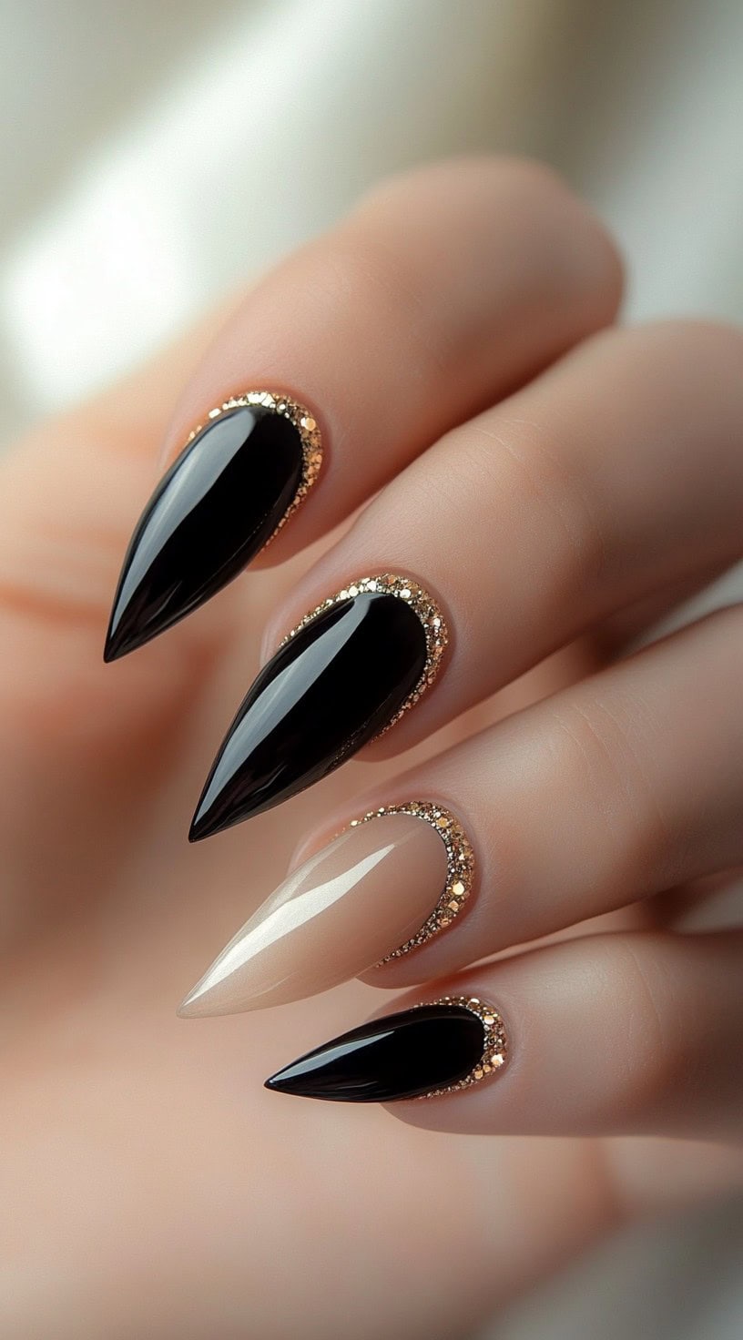 A hand with stiletto-shaped nails painted glossy black, featuring gold glitter accents along the cuticle area, with one accent nail in sheer nude.