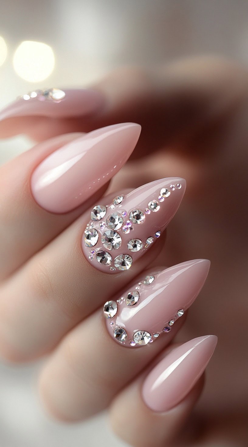 A close-up of long almond-shaped nails painted in a soft blush pink, with some nails adorned with sparkling clear crystals in varying sizes.