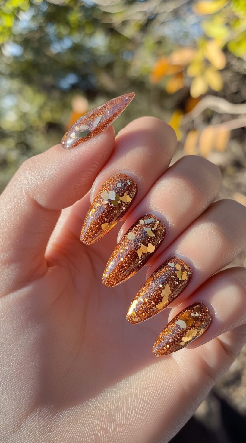 Hand with long almond-shaped nails covered in bronze glitter and chunky gold foil pieces.