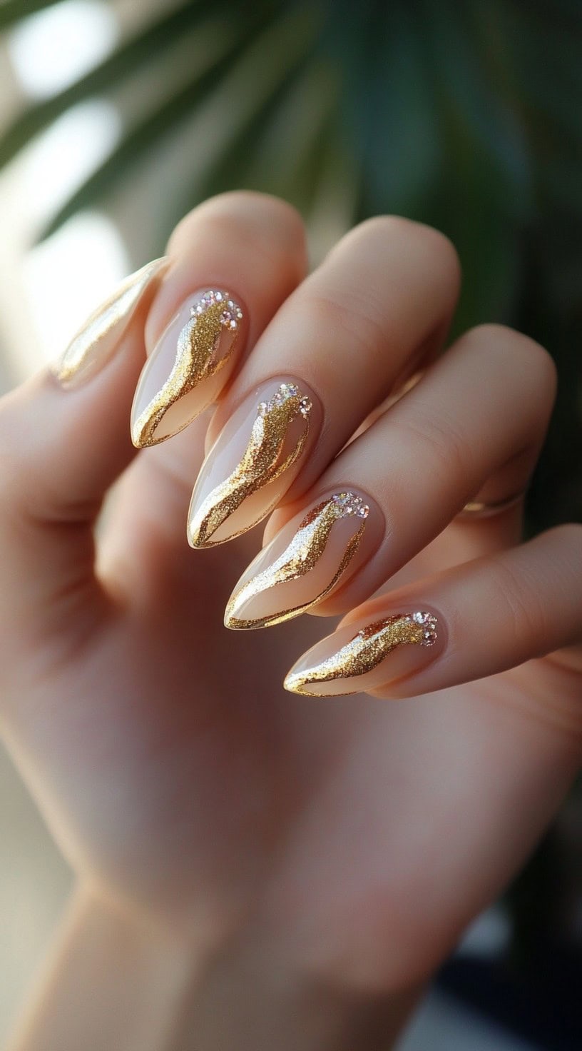 Hand with almond-shaped clear nails featuring gold glitter flame designs and small crystal accents.