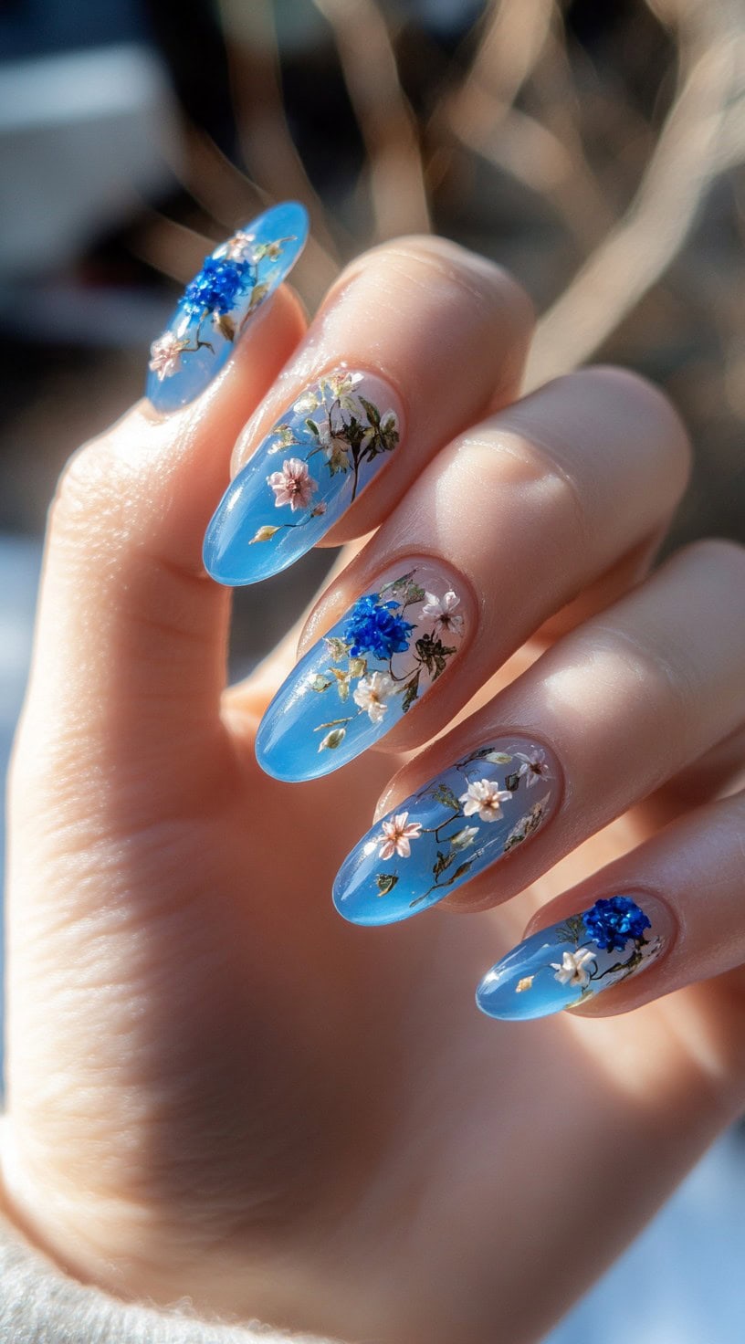 Clear blue oval nails with realistic floral designs in shades of white, pink, and blue.