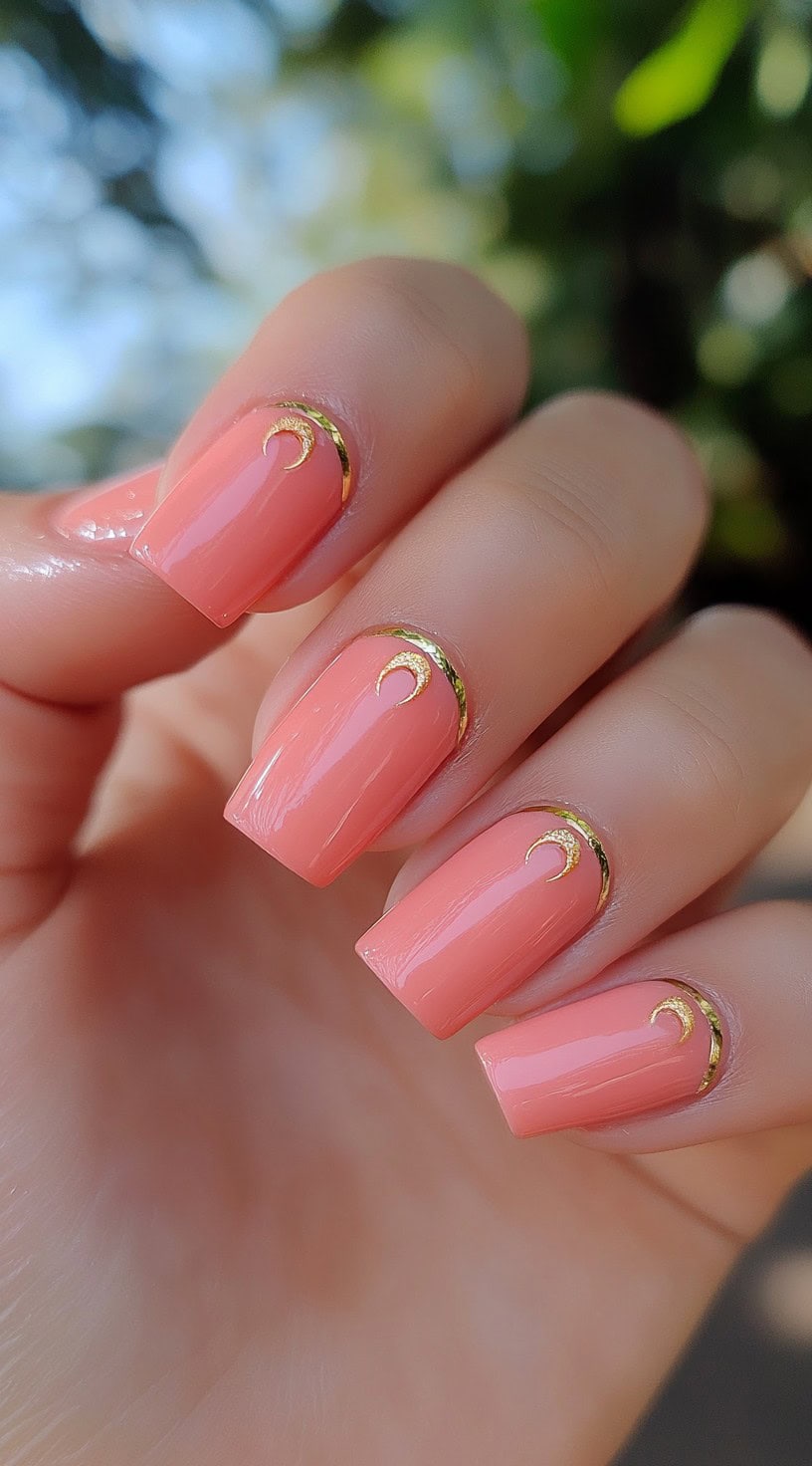 A hand with square nails painted coral pink, each featuring a delicate crescent moon-shaped gold detail near the cuticle.