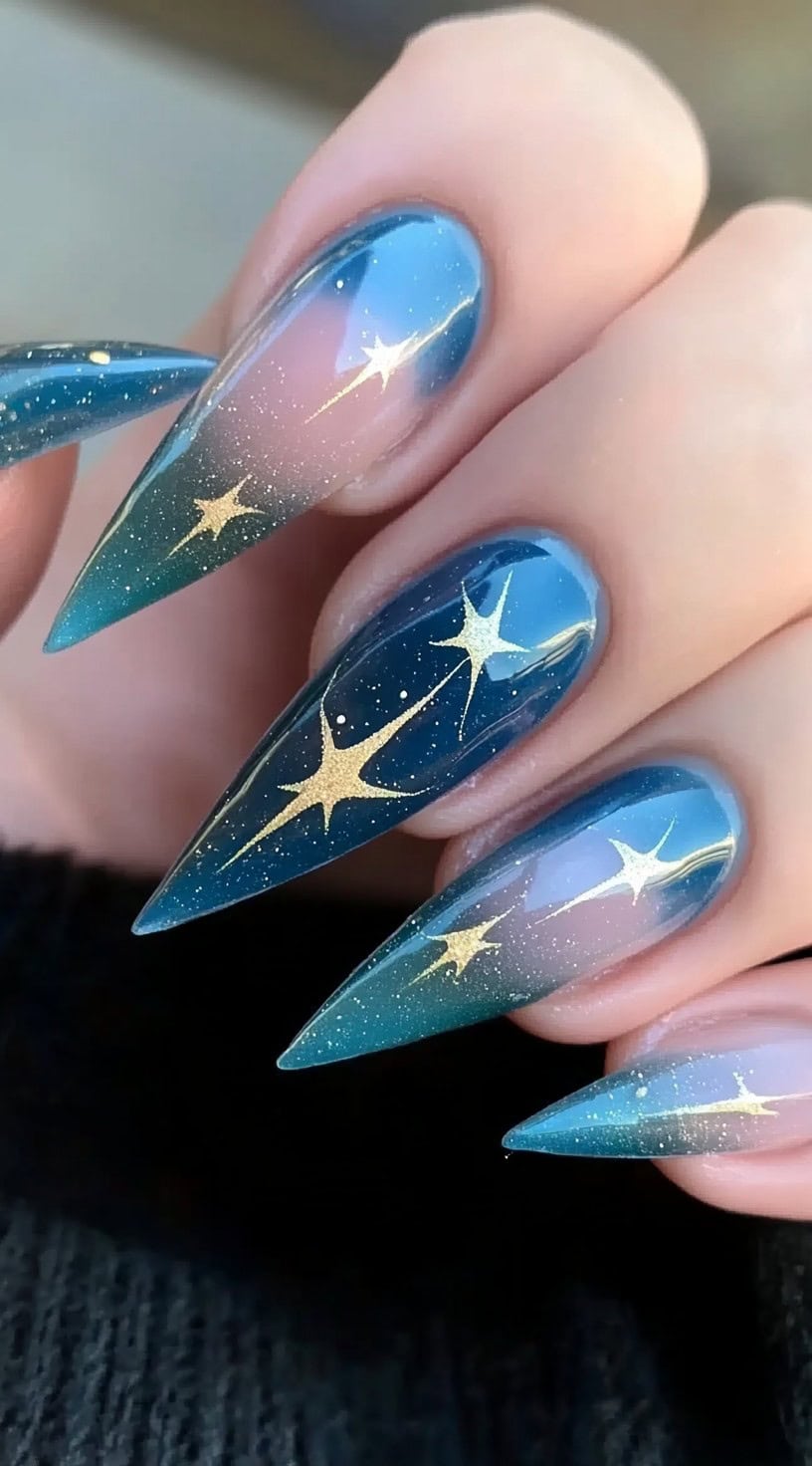 A set of stiletto nails featuring a deep blue aura gradient with golden star accents and shimmering specks of glitter.