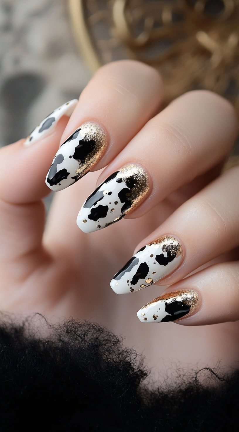 Almond-shaped nails featuring a classic black-and-white cow print design with gold glitter accents near the cuticles.