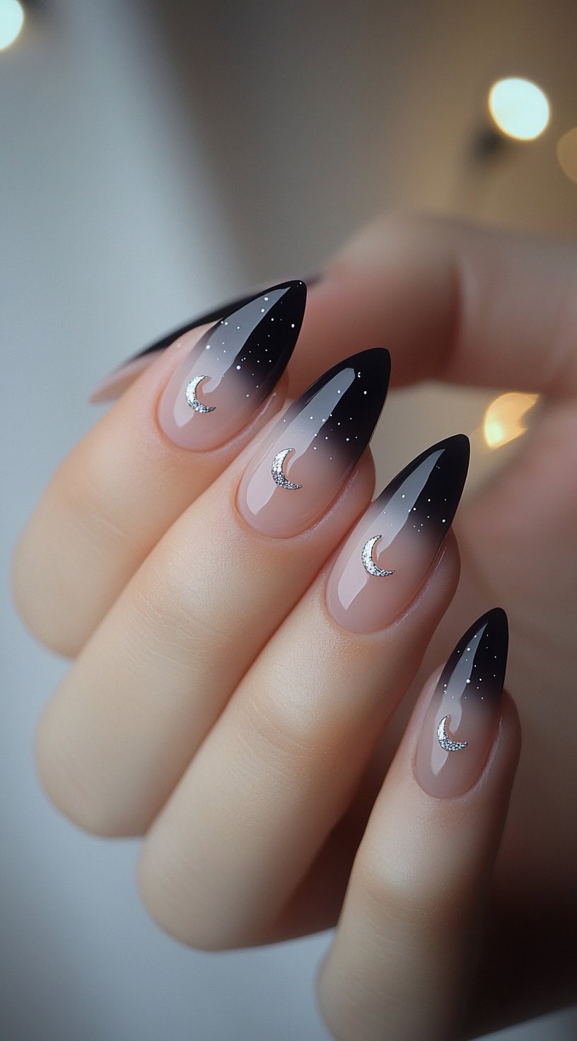 Stiletto nails showing a soft nude to black ombre with tiny stars and metallic crescent moon charms centered on each nail.