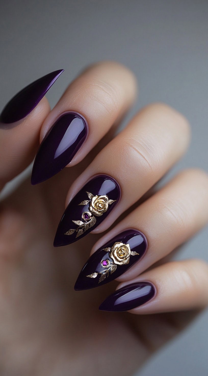 Deep plum almond nails with 3D gold rose charms and tiny crystals on the accent fingers.