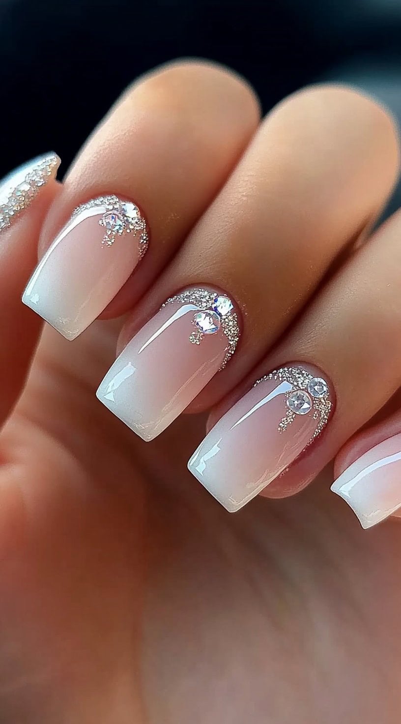A set of square nails featuring a soft pink-to-white ombre, adorned with silver glitter and tiny rhinestone embellishments along the cuticle.