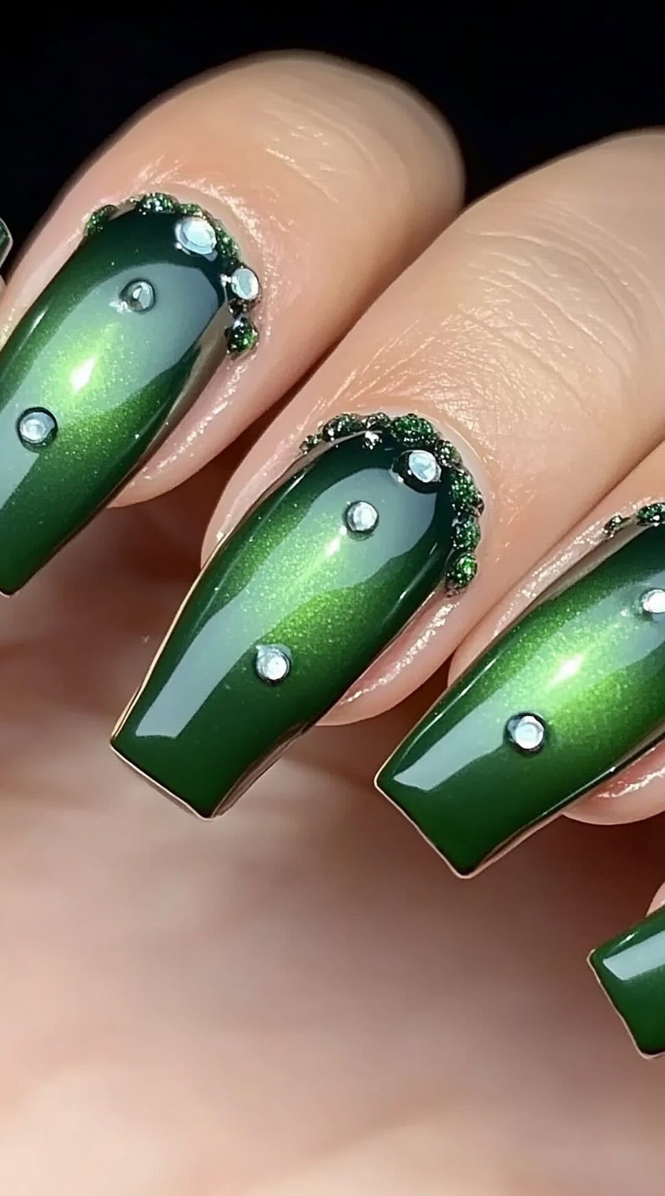 Coffin-shaped nails with a glossy emerald green finish, embellished with sparkling gemstones and textured rhinestone details near the cuticles.