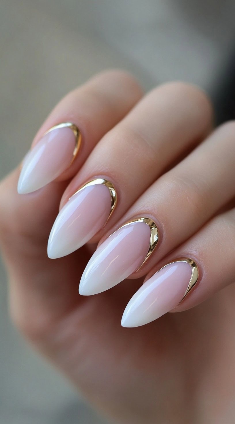 A close-up of almond-shaped nails featuring a soft pink-to-white ombré effect, enhanced by metallic gold accents along the cuticles.