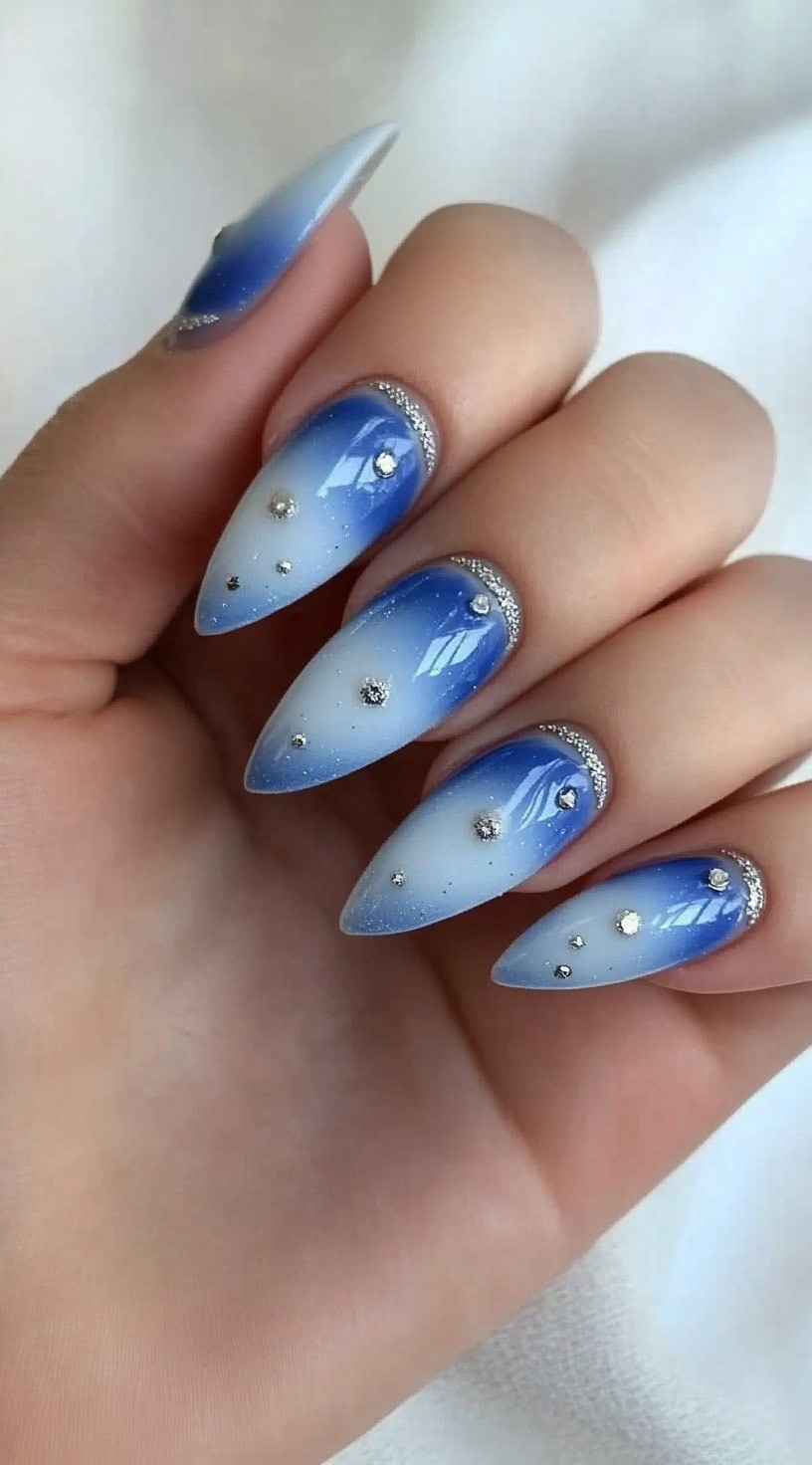 Almond-shaped nails featuring a soft blue aura ombré fading into icy white, adorned with sparkling crystals and glitter accents.