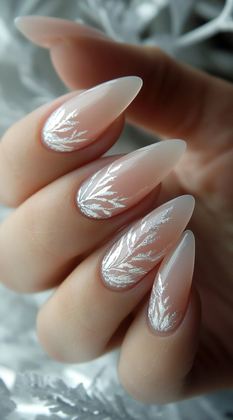 A close-up of long almond-shaped nails with a sheer nude base and delicate silver leaf-like designs at the cuticle.