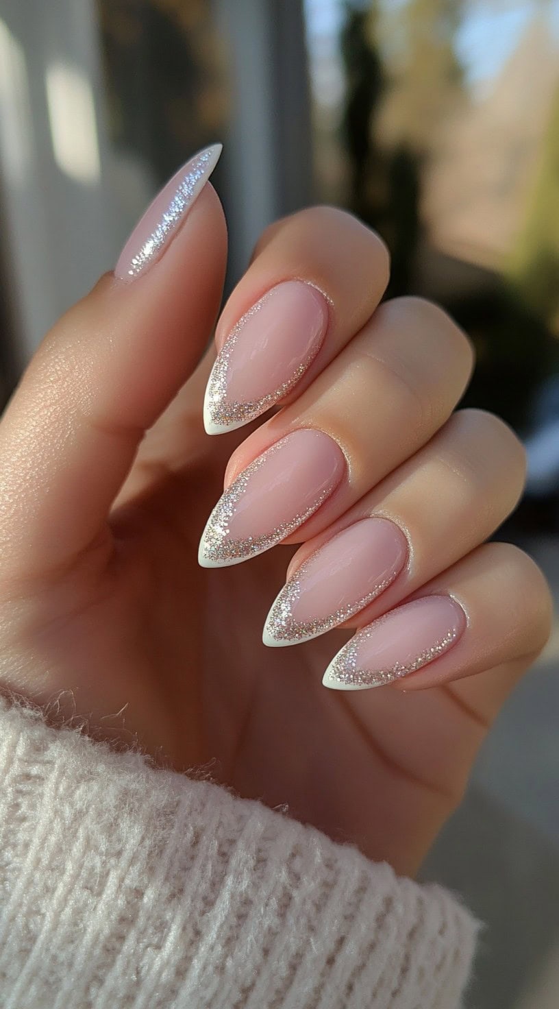 A hand with almond-shaped nails featuring a soft pink base and fine silver glitter French tips.