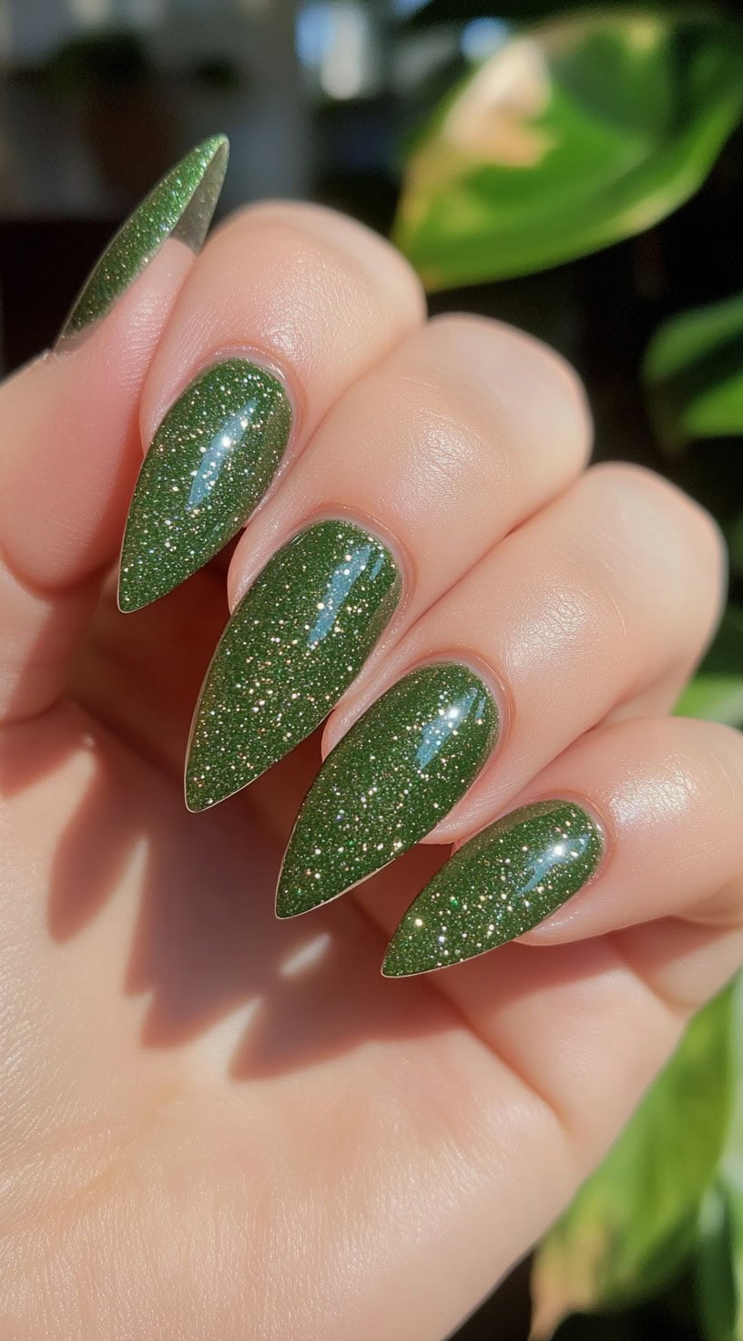 Pointed stiletto nails painted in a sparkly, dark matcha green polish that shimmers under light.