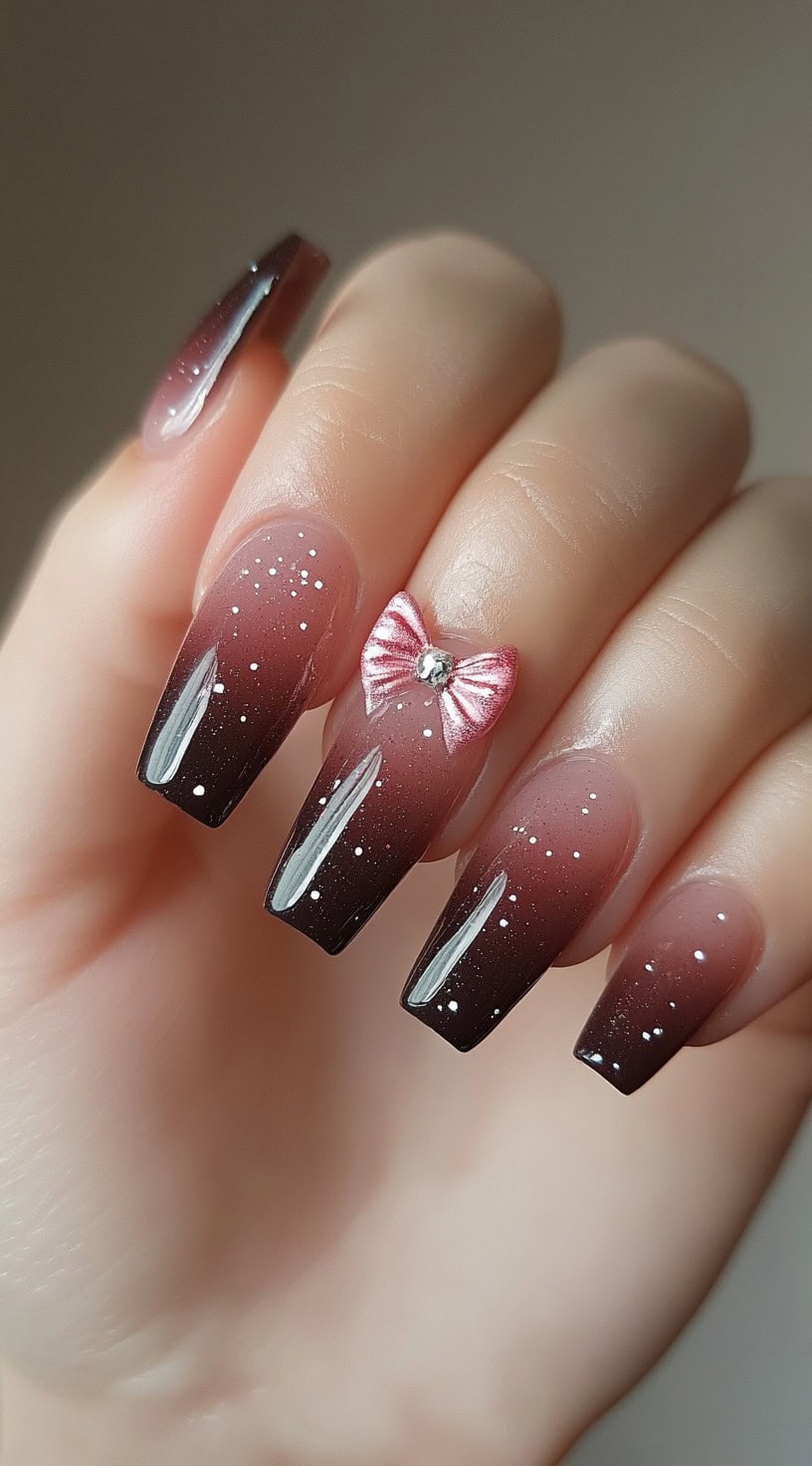 Long coffin nails with a brown to pink ombre gradient, white glitter specks, and a pink bow charm on the ring finger.