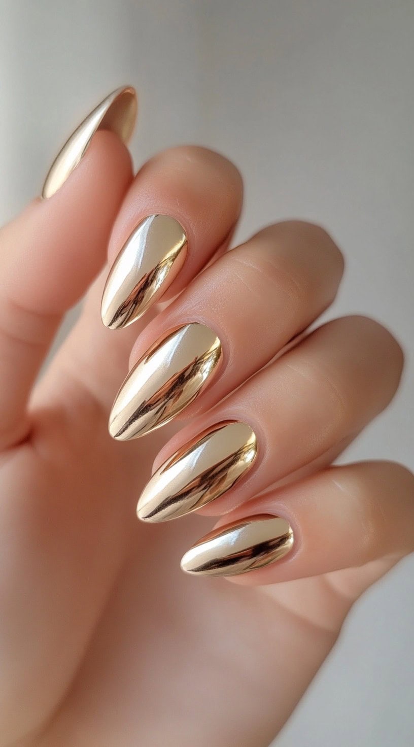 A close-up of almond-shaped nails coated in a high-shine gold chrome polish, reflecting light like liquid metal.